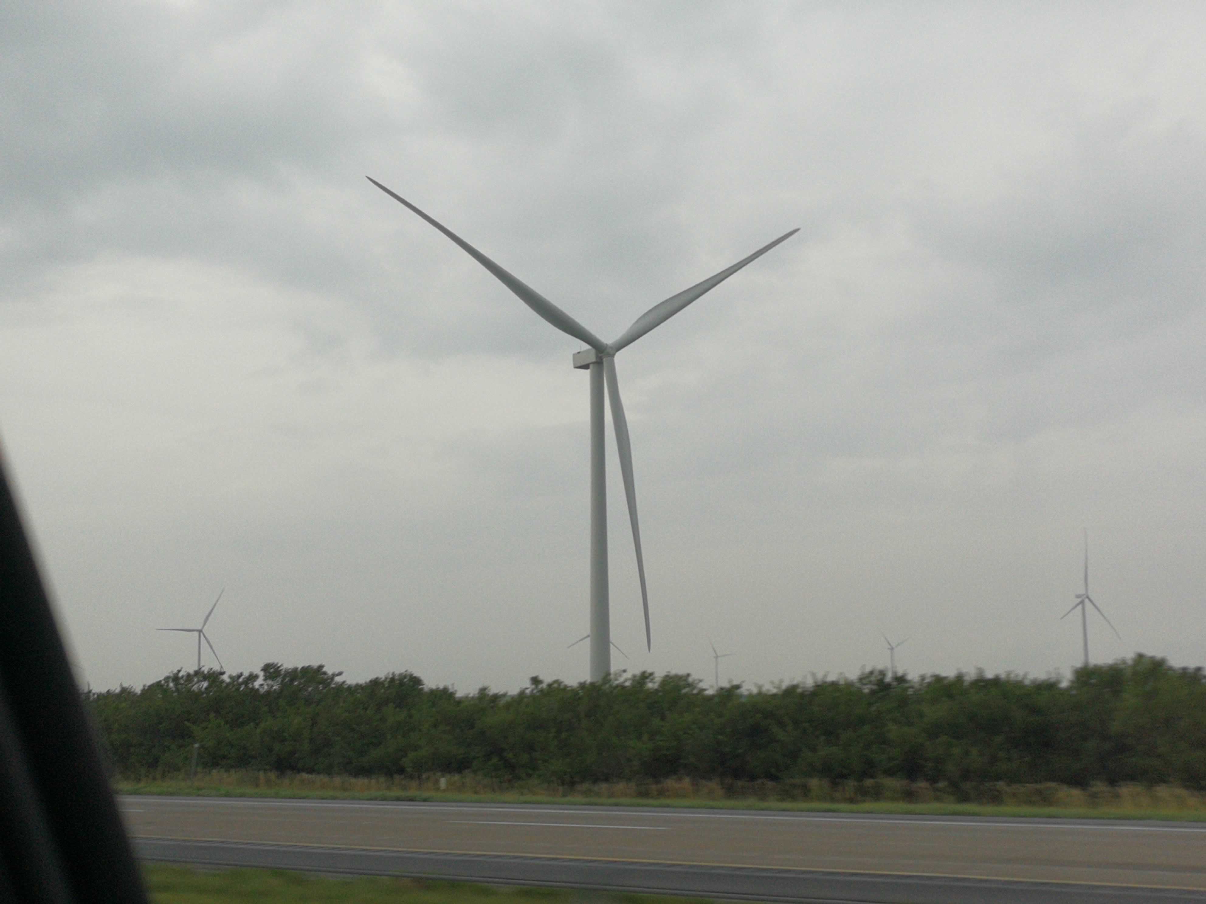 One wind turbine close by stands in the foreground of a field full of wind turbines. It is lightly raining.