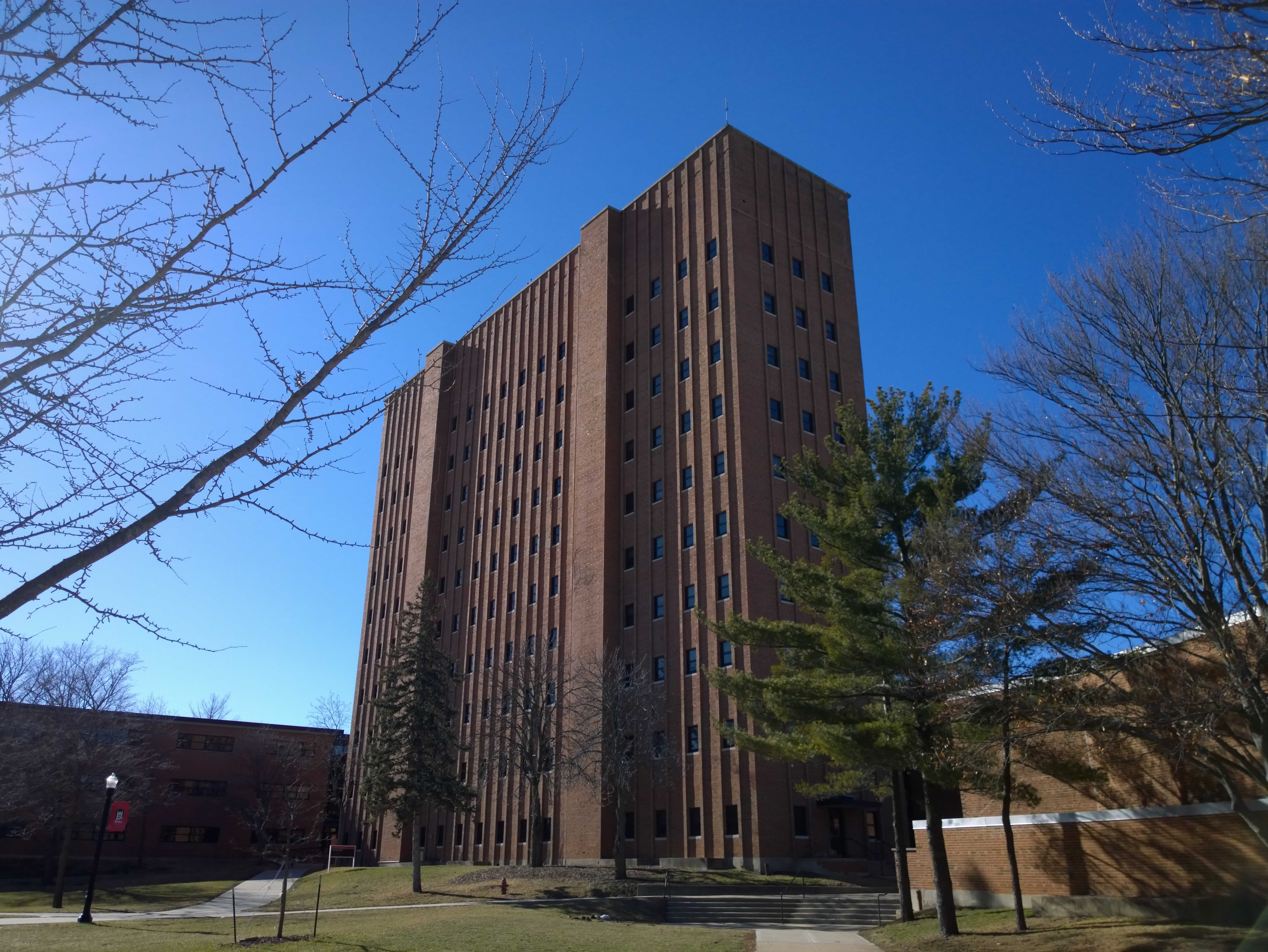 A building shaped similarly to a cereal box, made of brick with light ridges, rises up out of a grassy park, reaching into a cloudless blue sky.  Tree branches in the foreground form a frame around the building.
