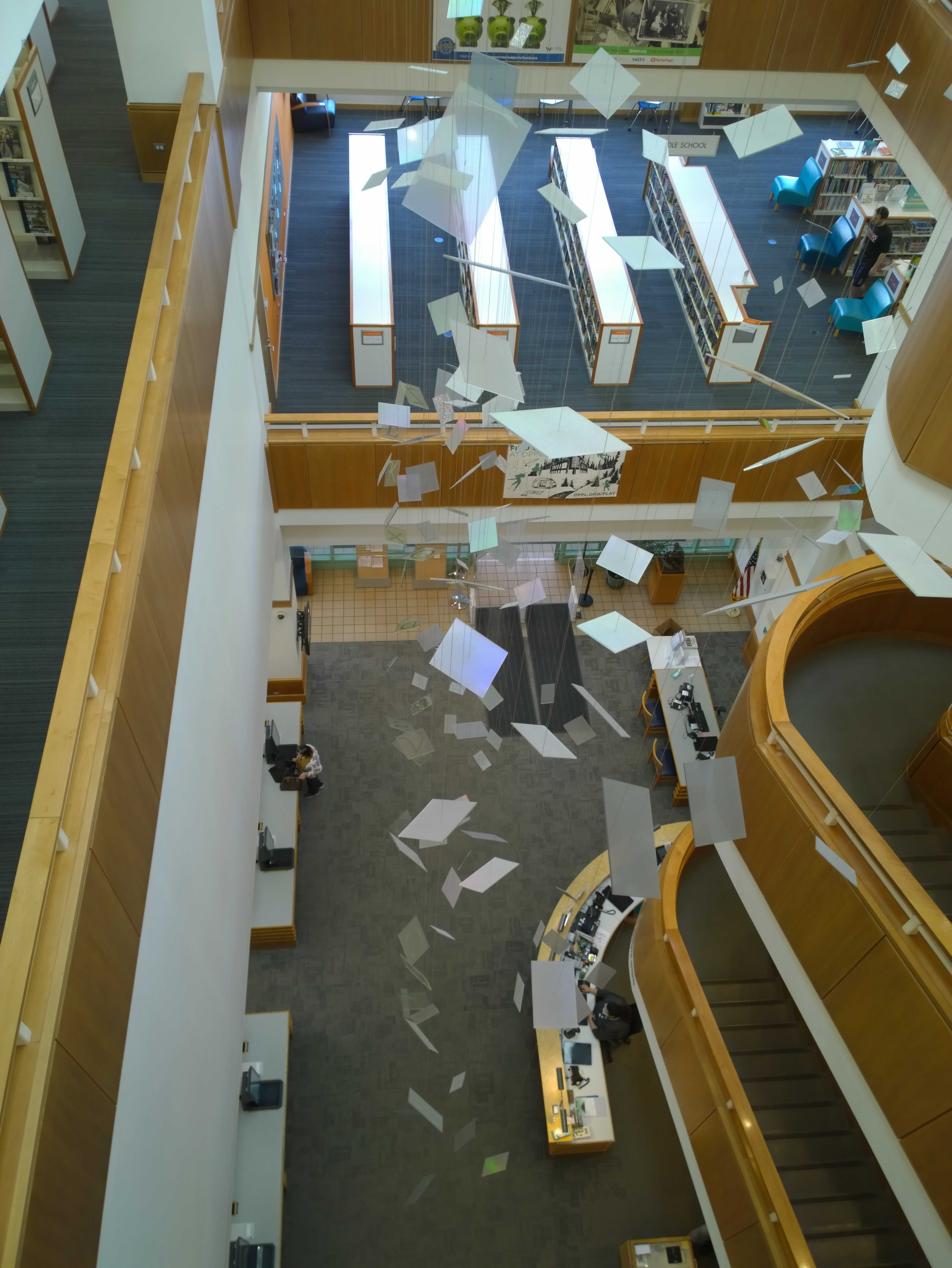 A bunch of index cards are hanging from the ceiling of a three story building. The photograph is taken from the third floor, showing the lower levels of the building with the index cards over top of them.