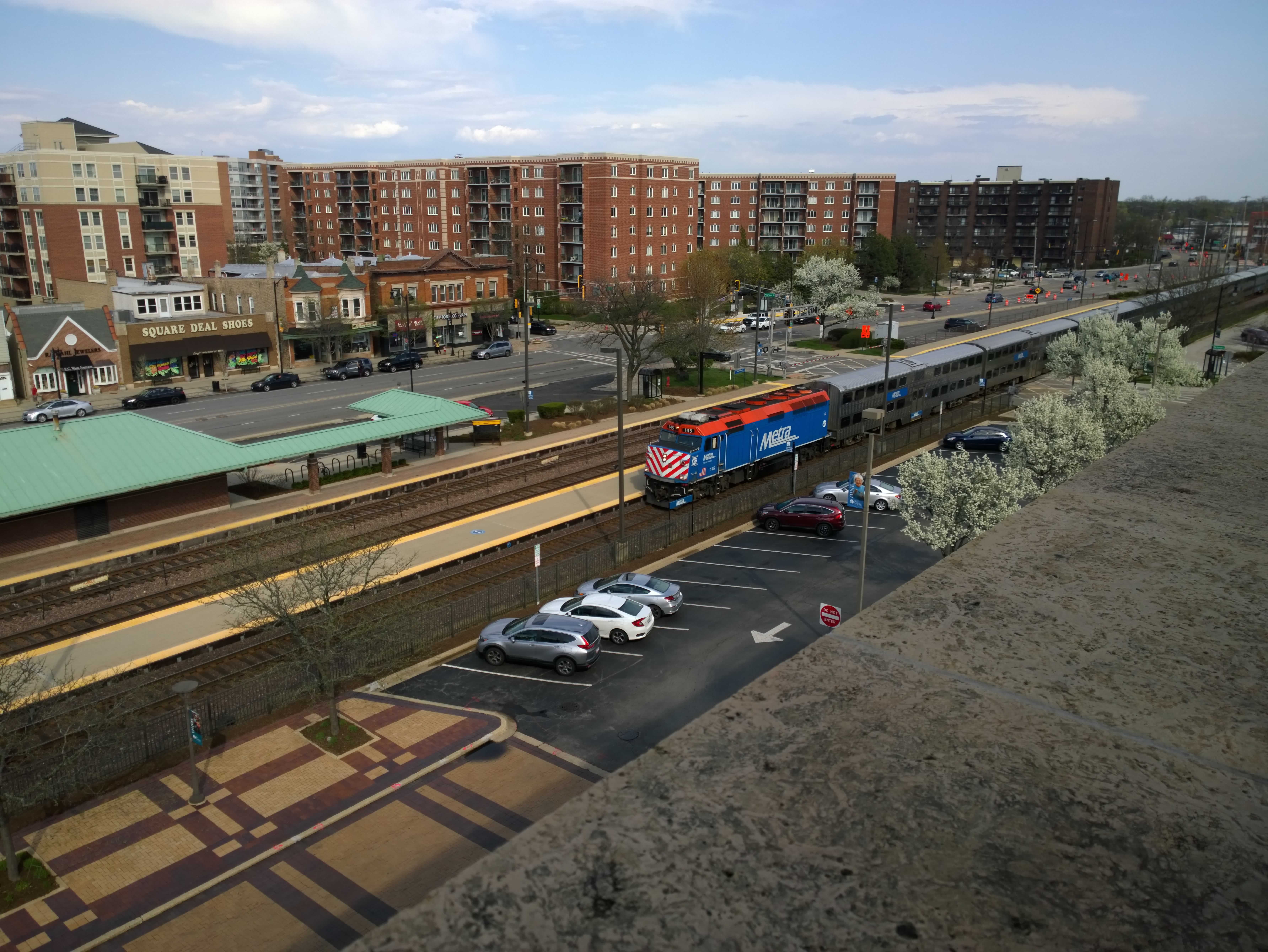 A blue and orange Metra engine leads seven passenger carriages down the tracks.  Trees in fresh bloom frame the left, while the green roof of the train station frames the left.