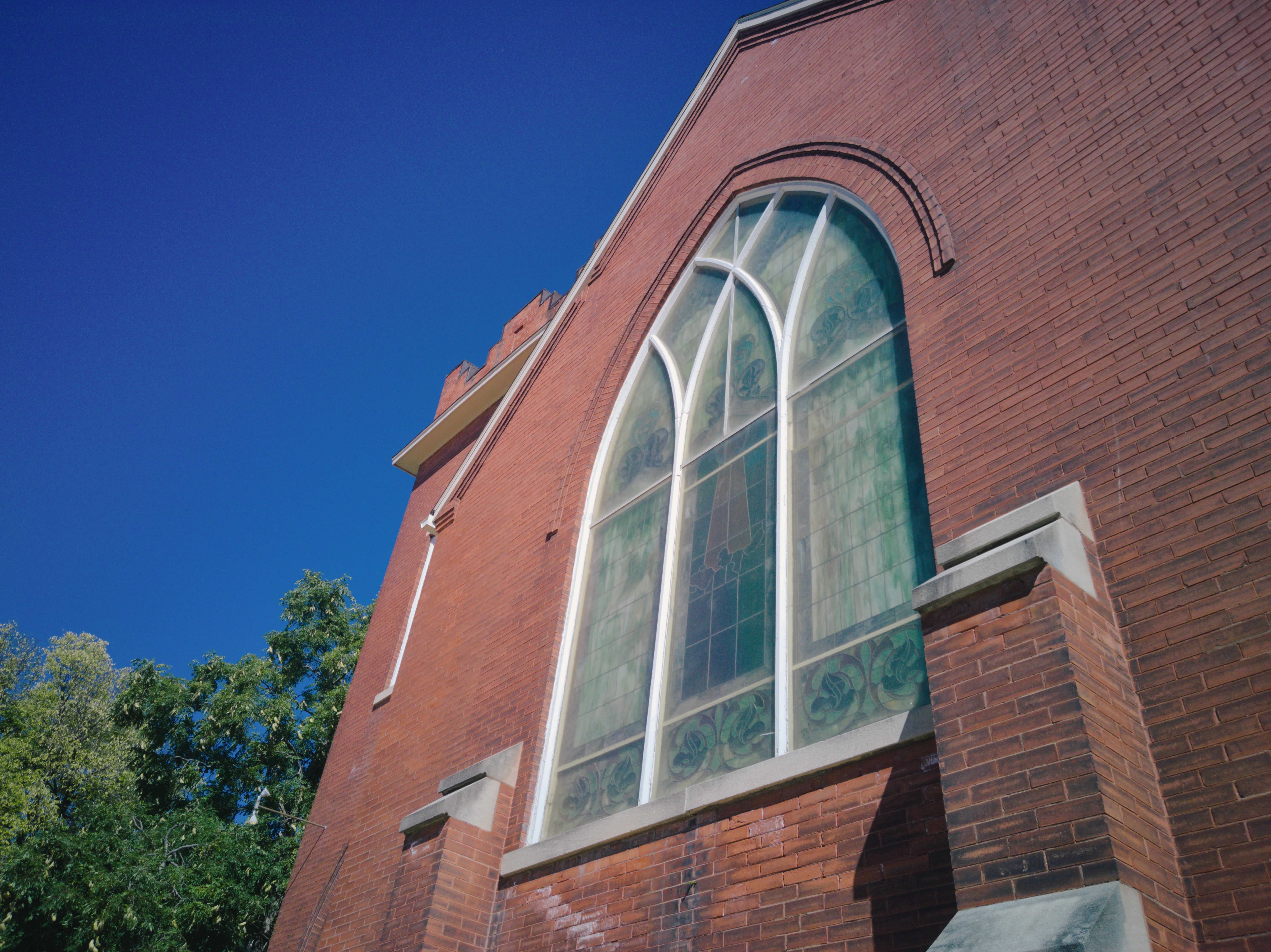 A brick church beneath a cloudless blue sky. A pale stained glass window arches upwards, sitting behind a protective green-ish layer.