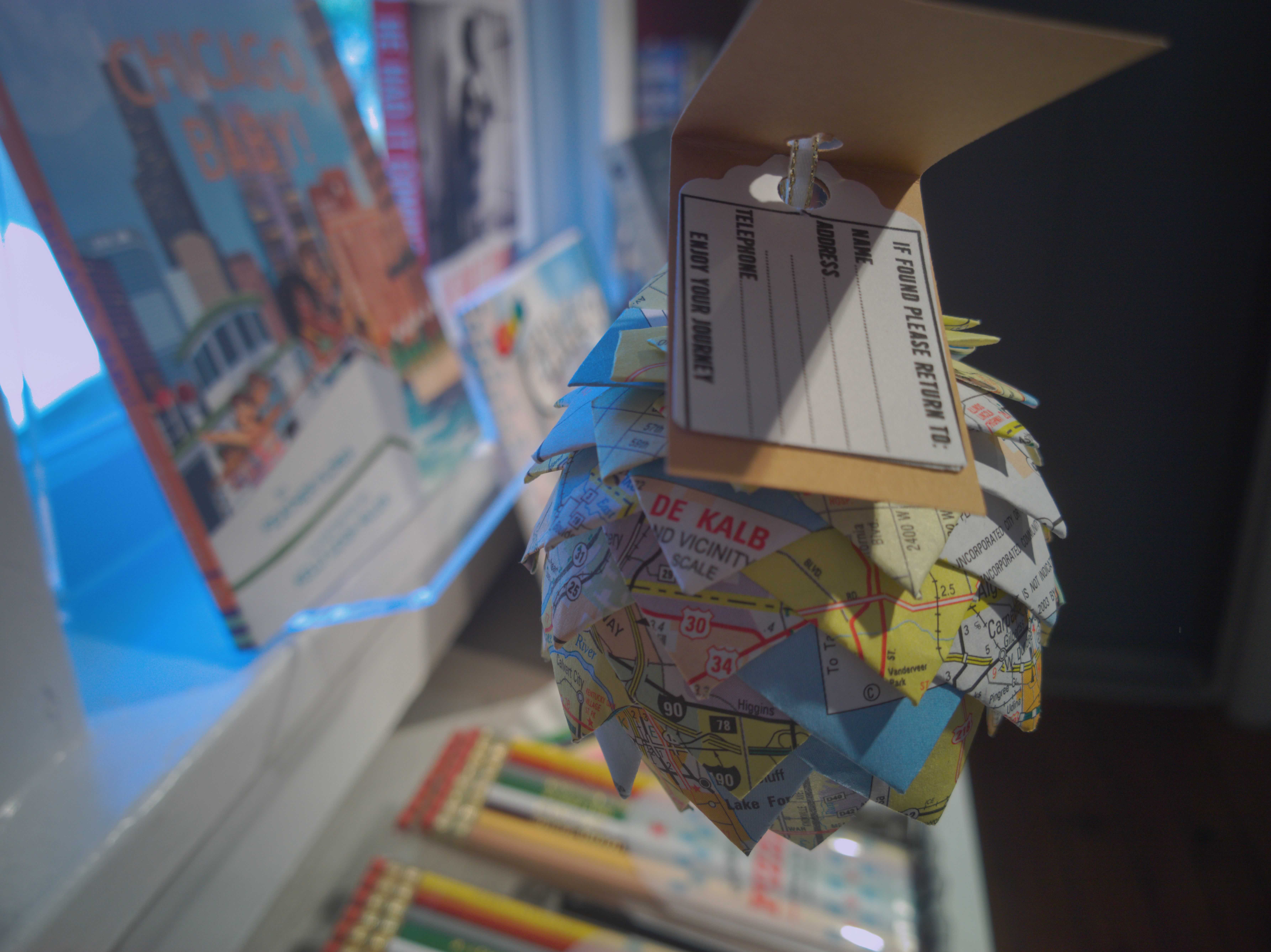 A papercraft pinecone made from a map sits against a bokeh blur background.  The presence of books and pencils on the shelves below implies a bookstore setting.