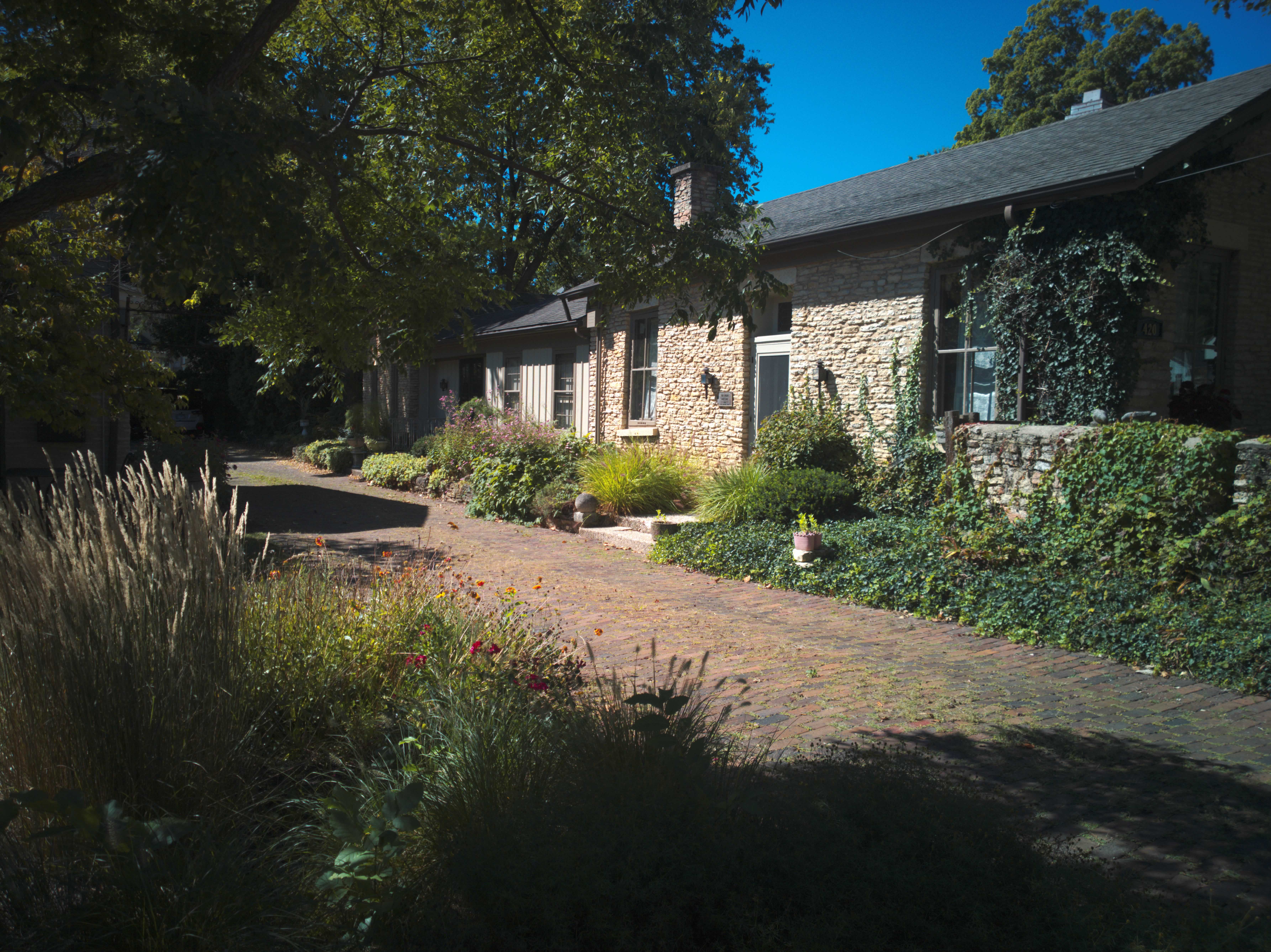 A tan brick house with overgrown ivy and shrubs sits alongside a brick driveway.